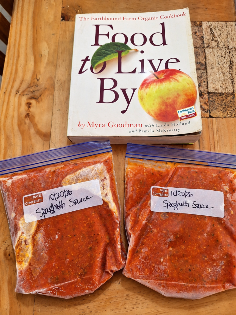 Two labeled freezer bags of homemade marinara sauce beside the Food to Live By cookbook on a wooden table.
