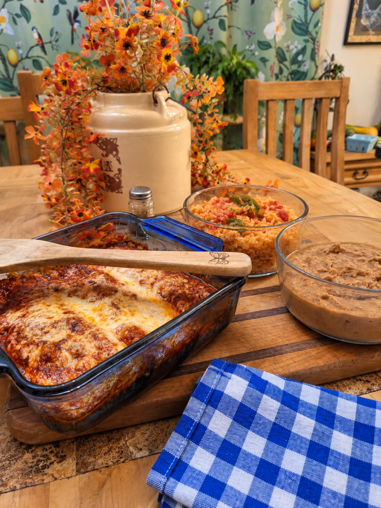 Homemade enchiladas topped with melted cheese and red enchilada sauce in a glass baking dish, served with Mexican rice and refried beans on a rustic farmhouse table.