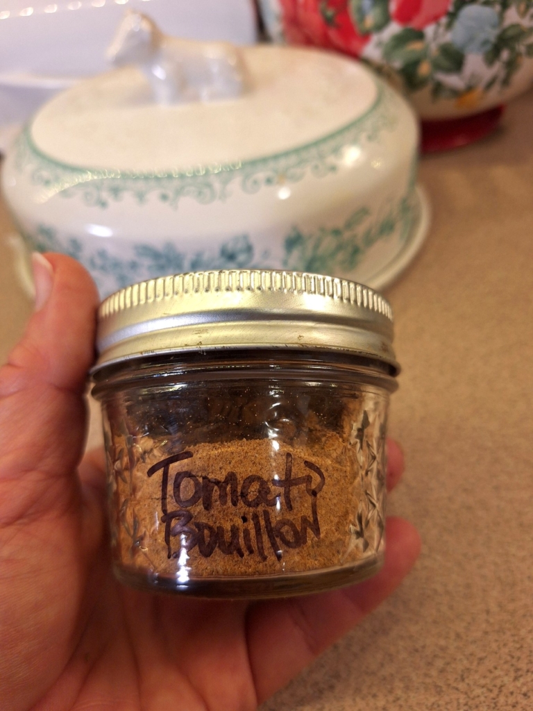 Small glass mason jar filled with homemade vegetarian tomato bouillon powder, with a handwritten label and silver lid, held in a farmhouse kitchen.